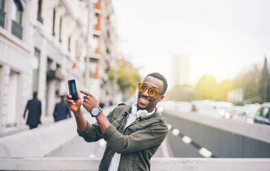 Glücklicher Mann mit Sonnenbrille, der sein Smartphone zeigt, während er in der Stadt steht - OCMF01588