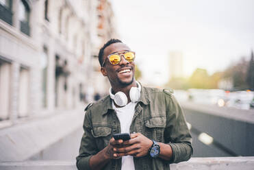 Fröhlicher Mann mit Sonnenbrille, der ein Smartphone benutzt, während er in der Stadt steht - OCMF01589