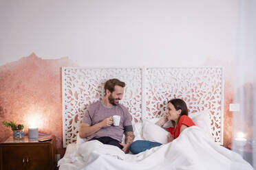 Lächelnder Mann mit Kaffee in der Hand im Gespräch mit seiner Frau, während er sich zu Hause auf dem Bett entspannt - EIF00142