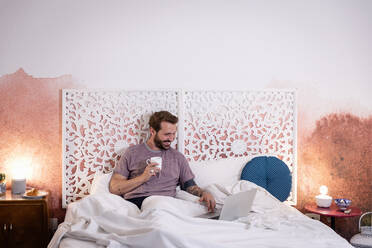 Smiling mid adult man holding coffee using laptop while relaxing on bed at home - EIF00151