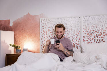 Lächelnder Mann, der einen Kaffeebecher hält und sein Smartphone benutzt, während er sich zu Hause auf dem Bett entspannt - EIF00153
