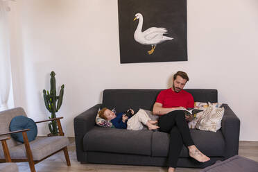 Mann liest eine Zeitschrift, während sein Sohn zu Hause auf dem Sofa mit einem Joystick spielt - EIF00163