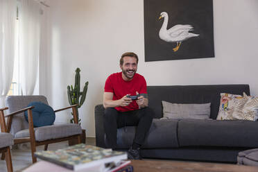 Fröhlicher erwachsener Mann spielt ein Videospiel, während er zu Hause auf dem Sofa sitzt - EIF00164
