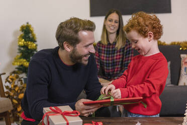 Lächelnde Frau betrachtet den Vater, der seinem Sohn im Wohnzimmer ein Weihnachtsgeschenk macht - EIF00168