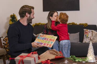 Man holding gift looking at son kissing mother in living room at home during Christmas - EIF00170