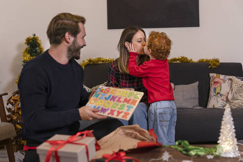 Mann, der ein Geschenk hält und seinen Sohn betrachtet, der seine Mutter zu Hause im Wohnzimmer küsst, während Weihnachten - EIF00170