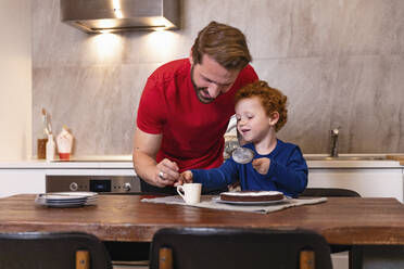 Glücklicher Vater und Sohn beim Verzieren von Schokoladenkuchen auf dem Esstisch in der Küche - EIF00183