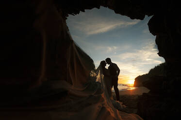 Side view of newlyweds hugging in dark cave on sand coast near water and blue sky at sunset - ADSF07235