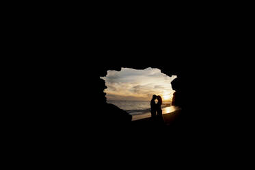 Side view of newlyweds hugging in dark cave on sand coast near water and blue sky at sunset - ADSF07236