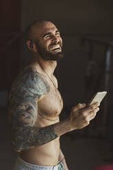 Seitenansicht eines gutaussehenden, tätowierten Mannes mit Smartphone, der lacht und Musik hört, während er in einem modernen Fitnessstudio beim Training steht - ADSF07237