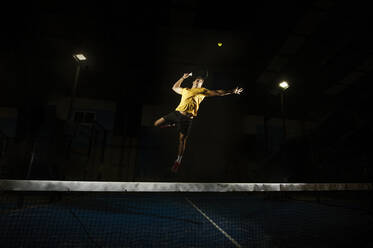 Sportlicher Mann, der einen Tennisschläger hält und springt, um einen Ball auf einem Tennisplatz im Dunkeln zu treten - ADSF07239