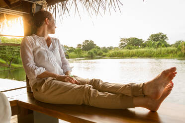 Seitenansicht einer barfüßigen Frau, die in einem Pavillon sitzt und auf einen ruhigen Teich an einem sonnigen Tag in Sri Lanka blickt - ADSF07243