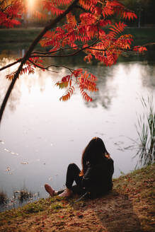Junge Frau betrachtet die Aussicht, während sie im Herbst am See sitzt - CAVF87679