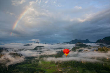 Ein Sonnenaufgang über Vang Vieng, mit einem Regenbogen über den Wolken, während im Vordergrund ein Heißluftballon aufsteigt, Laos, Indochina, Südostasien, Asien - RHPLF17154