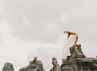 Seitenansicht einer nicht erkennbaren Frau in BH und elegantem Rock, die sich nach hinten beugt, während sie auf einem Felsen vor einem wolkenverhangenen grauen Himmel steht - ADSF07257