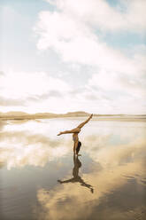 Side view of anonymous female in swimwear performing handstand on wet sand near sea against cloudy sky - ADSF07259