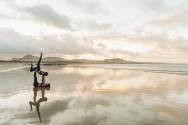 Side view of two barefoot females in sportswear performing exercise on wet sand near sea on overcast day - ADSF07263