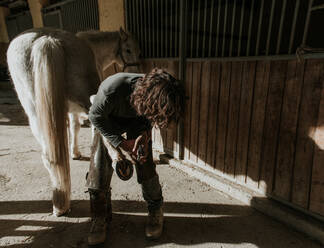 Side view of adult blacksmith using hammer to put horseshoe on hoof of horse near stable on ranch - ADSF07264