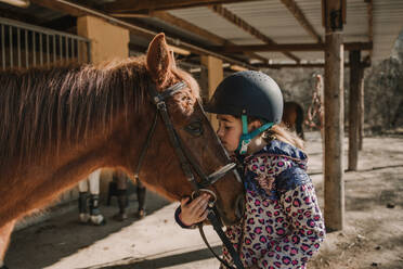 Niedliches kleines Mädchen mit Helm, das ein weißes Pferd küsst, während es in der Nähe von Stallungen während des Reitunterrichts auf einer Ranch steht - ADSF07281