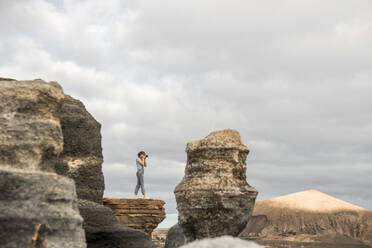 Seitenansicht einer nicht erkennbaren Frau, die Fotos von einer schönen Landschaft macht, während sie inmitten von rauen Steinsäulen gegen einen bedeckten Himmel steht - ADSF07287