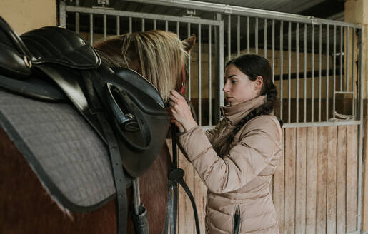 Seitenansicht einer jungen Frau mit Helm, die ein wunderschönes Pferd in der Nähe des Stalls auf einer Ranch sattelt - ADSF07297