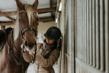 Seitenansicht einer jungen Frau mit Helm, die ein wunderschönes Pferd in der Nähe des Stalls auf einer Ranch sattelt - ADSF07298