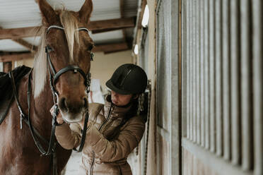 Side view of young female in helmet putting saddle on wonderful horse near stable on ranch - ADSF07298