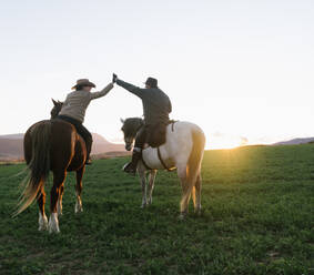 Rückenansicht eines Mannes und einer Frau, die auf Pferden reiten und sich gegenseitig vor dem Hintergrund des Sonnenuntergangs auf einer Ranch 