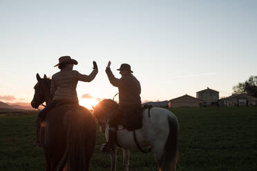 Rückenansicht eines Mannes und einer Frau, die auf Pferden reiten und sich gegenseitig vor dem Hintergrund des Sonnenuntergangs auf einer Ranch 