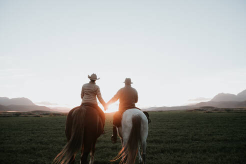 Rückenansicht eines Mannes und einer Frau, die auf Pferden reiten und sich an den Händen halten, bei Sonnenuntergang auf einer Ranch - ADSF07305