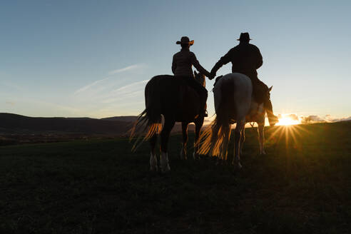 Rückenansicht eines Mannes und einer Frau, die auf Pferden reiten und sich an den Händen halten, bei Sonnenuntergang auf einer Ranch - ADSF07306