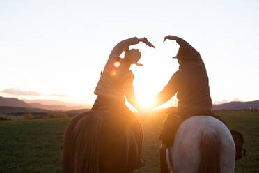 Rückenansicht eines Mannes und einer Frau, die auf Pferden reiten und mit den Händen ein Herz bilden, vor dem Hintergrund des Sonnenuntergangs auf einer Ranch - ADSF07307