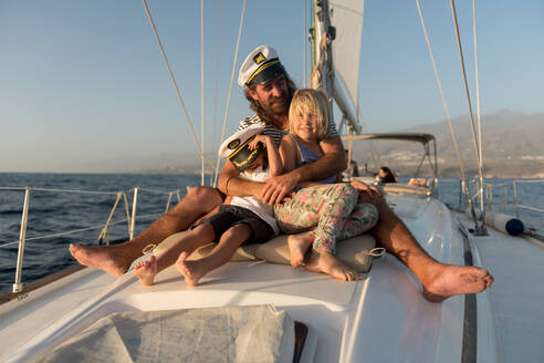 Positiver Vater umarmt glückliche Kinder in Kapitänsmützen und sitzt an Deck eines teuren Bootes, das an einem sonnigen Tag auf dem Wasser schwimmt - ADSF07319