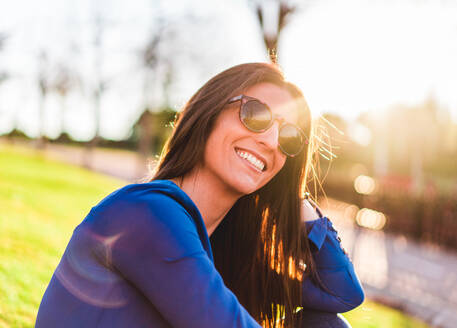 Porträt einer jungen Frau mit Sonnenbrille im Gras sitzend im Park - ADSF07322