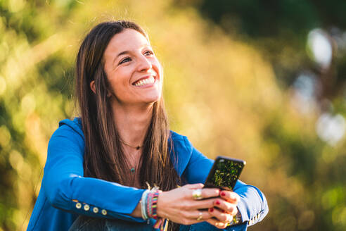 Porträt einer jungen Frau, die ein Smartphone benutzt und im Gras eines Parks sitzt - ADSF07323
