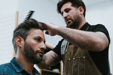 From below barber combing hair of handsome stylish male sitting in barbershop - ADSF07331