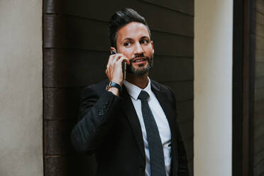 Erwachsener gutaussehender eleganter Geschäftsmann in formellem Anzug, der wegschaut und mit einem Mobiltelefon in der Nähe der Wand spricht - ADSF07336