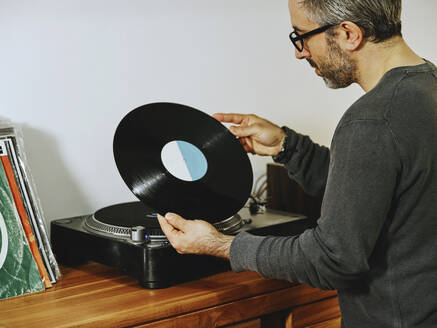 Seitenansicht eines seriösen Mannes, der einen Plattenspieler einstellt und Lieder auf einer Retro-Vinylscheibe genießt, während er zu Hause chillt - ADSF07432