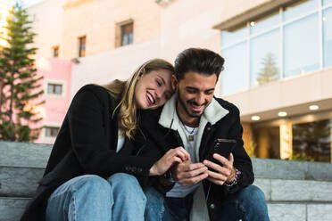 Happy young couple in stylish clothes sharing earphones and enjoying music on smartphone while sitting together on stairs near city building - ADSF07459