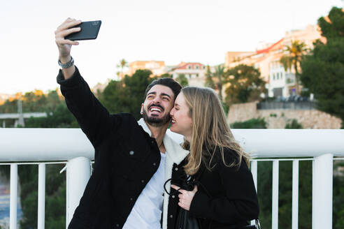 Fröhliches junges gemischtrassiges Paar in Freizeitkleidung, das ein Selfie mit dem Handy macht, während es zusammen auf einer Brücke mit grünen Bäumen und städtischen Gebäuden im Hintergrund steht - ADSF07460