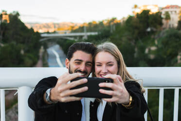 Fröhliches junges gemischtrassiges Paar in Freizeitkleidung, das ein Selfie mit dem Handy macht, während es zusammen auf einer Brücke mit grünen Bäumen und städtischen Gebäuden im Hintergrund steht - ADSF07461