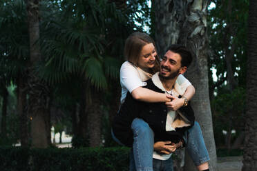Happy ethnic man in stylish jacket and jeans giving piggyback ride to cheerful girlfriend while spending time together in green park with tropical plants - ADSF07464