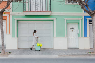Side view of young stylish female with yellow balloons and suitcase walking on city street next to old styled colorful building - ADSF07465