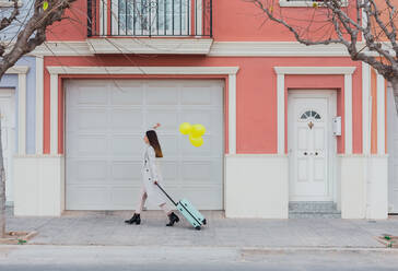 Seitenansicht der jungen stilvollen Frau mit gelben Luftballons und Koffer zu Fuß auf der Straße neben alten stilisierten bunten Gebäude - ADSF07466