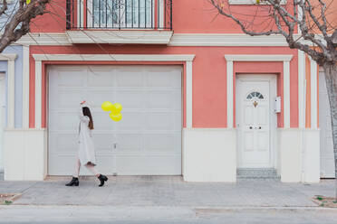 Side view of unrecognizable stylish female covering face with yellow balloons walking with suitcase on city street next to old styled colorful building - ADSF07467