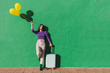Moderne, modische, sorglose Frau, die ein Bündel gelber Luftballons und einen Koffer hält, während sie an einer grünen Wand in einem sonnigen Tag steht - ADSF07468