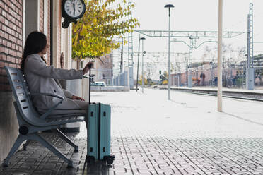 Seitenansicht von jungen stilvollen weiblichen Reisenden mit Koffer sitzen auf Bank und warten auf den Transport am Bahnhof in sonnigen Tag - ADSF07471