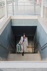 From above of elegant stylish young female traveler with suitcase walking up stairs at train station - ADSF07472