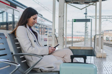 Seitenansicht einer modernen Reisenden mit Koffer, die auf einer Bank auf dem Bahnsteig sitzt und ihr Smartphone benutzt, während sie auf den Zug wartet - ADSF07475