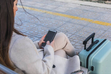 Von oben aufgenommene unerkennbare weibliche Reisende mit Koffer, die auf einer Bank auf dem Bahnsteig eines Bahnhofs sitzt und ihr Smartphone benutzt, während sie auf den Zug wartet - ADSF07476